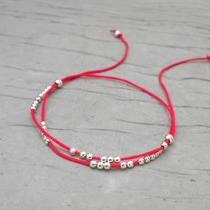 Peut inclure: Un bracelet rouge avec deux rangées parallèles. De petites perles argentées brillantes sont enfilées sur le cordon rouge. Le bracelet a des nœuds coulissants réglables. Le fond est une surface grise texturée.