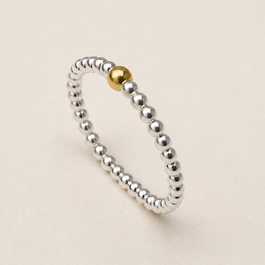 Peut inclure: Une bague composée d'un anneau continu de petites perles sphériques argentées polies, avec une seule perle sphérique dorée, lisse et légèrement plus grande, placée comme point focal dans la disposition circulaire de l'anneau.