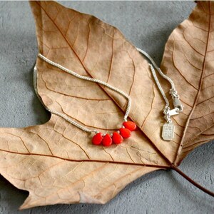 Puede incluir: Un collar de cadena de plata con cinco peque&ntilde;as cuentas de color rojo brillante en forma de l&aacute;grima, dispuestas en un grupo en el centro. El collar incluye una fina cadena de plata y una peque&ntilde;a etiqueta met&aacute;lica rectangular cerca del cierre.