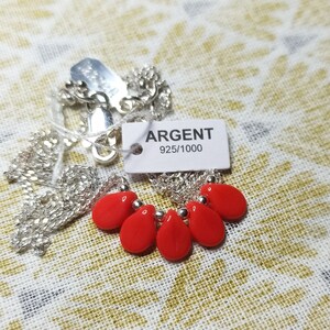 Puede incluir: Un collar de cadena plateada con cinco cuentas rojas brillantes en forma de l&aacute;grima separadas por peque&ntilde;as esferas plateadas. Una etiqueta rectangular blanca unida al collar muestra el texto ARGENT 925/1000, indicando el metal.