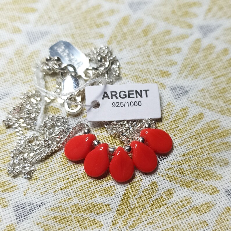 Puede incluir: Un collar de cadena plateada con cinco cuentas rojas brillantes en forma de l&aacute;grima separadas por peque&ntilde;as esferas plateadas. Una etiqueta rectangular blanca unida al collar muestra el texto ARGENT 925/1000, indicando el metal.