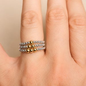 Könnte beinhalten: Ein stapelbares Ring-Set bestehend aus drei einzelnen Ringen aus kleinen silberfarbenen Perlen. In der Mitte des Stapels befindet sich eine dreieckige Anordnung aus sechs größeren, polierten goldfarbenen Perlen, die über den drei Ringen liegen.