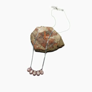 Puede incluir: Un collar de cadena plateada con cinco peque&ntilde;as piedras rosadas en forma de l&aacute;grima con una superficie texturizada y moteada. El collar est&aacute; colocado sobre una roca irregular de tonos marrones, beige y grises sobre un fondo blanco.