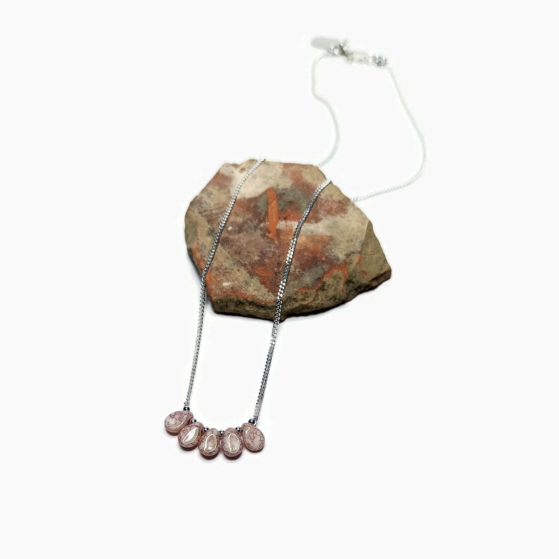 Puede incluir: Un collar de cadena plateada con cinco peque&ntilde;as piedras rosadas en forma de l&aacute;grima con una superficie texturizada y moteada. El collar est&aacute; colocado sobre una roca irregular de tonos marrones, beige y grises sobre un fondo blanco.