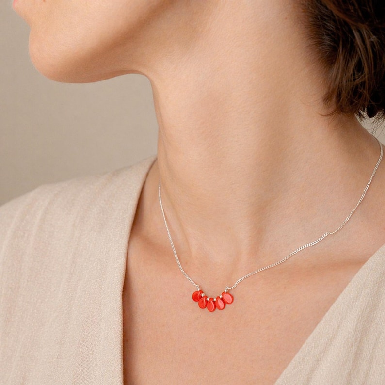 Puede incluir: Un collar de cadena plateada que presenta una disposici&oacute;n central de cinco peque&ntilde;os dijes lisos en forma de gota de color naranja brillante. Los dijes est&aacute;n espaciados uniformemente a lo largo de la delicada cadena de metal, creando un punto focal.