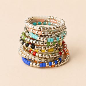 Peut inclure: Une pile de plusieurs bagues extensibles composées de perles argentées métalliques alternées avec des perles en plastique colorées. Les bagues présentent diverses couleurs dont blanc, pêche, turquoise, vert, noir, jaune, rouge, orange et bleu.