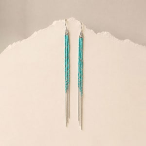Peut inclure: Une paire de longues boucles d'oreilles pendantes. Chaque boucle d'oreille est composée d'un crochet argenté et d'une section turquoise perlée, se transformant en plusieurs brins de chaîne argentée. Les boucles d'oreilles sont présentées sur un fond texturé blanc cassé.