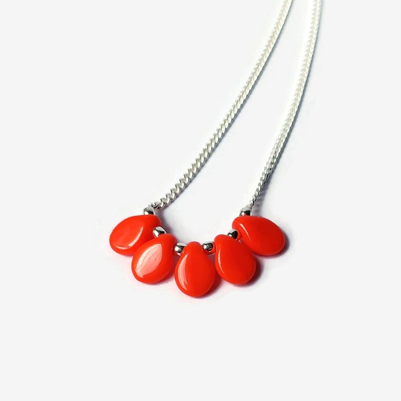 Puede incluir: Un collar con una cadena de tono plateado y cinco cuentas lisas en forma de l&aacute;grima de un color rojo anaranjado vibrante. Las cuentas est&aacute;n dispuestas en un grupo en el centro de la cadena, separadas por peque&ntilde;as cuentas espaciadoras met&aacute;licas.
