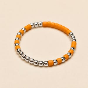 Könnte beinhalten: Ein kreisförmiger Stretch-Ring mit einem abwechselnden Muster aus kleinen, runden, metallisch silberfarbenen Perlen und matten orangefarbenen Perlen. Die Perlen sind in einer durchgehenden Schleife aufgereiht und bilden ein schlichtes Design.