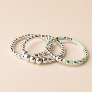 Peut inclure: Trois bracelets de perles. Deux bracelets sont argentés avec de petites perles rondes. Le troisième bracelet est vert clair avec des perles argentées. Les bracelets sont empilés sur une surface beige clair.