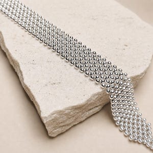 Może przedstawiać: Szeroka, elastyczna bransoletka lub naszyjnik typu choker, złożona z wielu rzędów małych, polerowanych, srebrzystych metalowych kulek połączonych w gęsty, jednolity wzór siatki, tworzący teksturowaną i odblaskową powierzchnię.