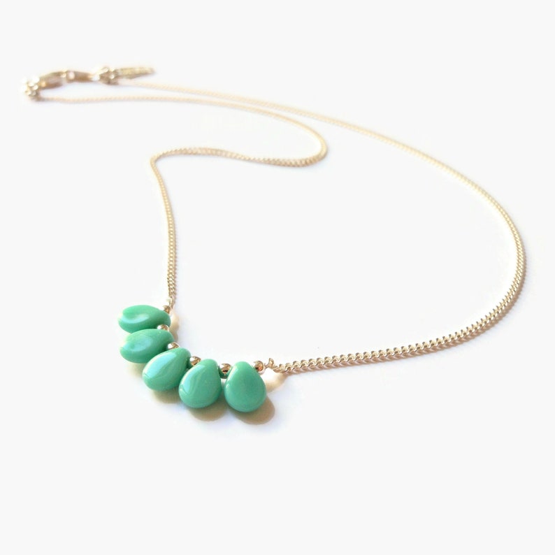 Puede incluir: Un collar de cadena en tono dorado con cinco cuentas lisas en forma de l&aacute;grima de color turquesa dispuestas en un grupo en el centro. La delicada cadena de metal incluye un peque&ntilde;o mecanismo de cierre en el extremo, con un dise&ntilde;o sencillo.