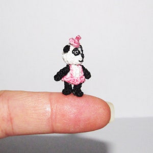 Puede incluir: Un pequeño panda de crochet con un vestido rosa y un lazo rosa. El panda está de pie sobre un dedo.