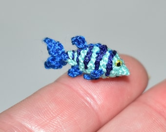 Puede incluir: Un pequeño pez tejido a ganchillo en tonos azules y turquesas, con rayas azul oscuro y un ojo amarillo. La criatura acuática hecha a mano descansa sobre un dedo, mostrando su pequeño tamaño y detalles intrincados. Un ejemplo encantador de artesanía en miniatura.