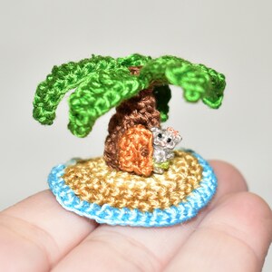 May include: A miniature crocheted island with a palm tree and a small animal sitting under the tree. The island is made of brown yarn and has a blue border. The palm tree is green and has a brown trunk. The animal is gray and has a white belly.