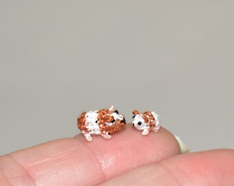 May include: Two small crocheted guinea pigs, one brown and white and one white, are sitting on a finger. The guinea pigs are facing each other.
