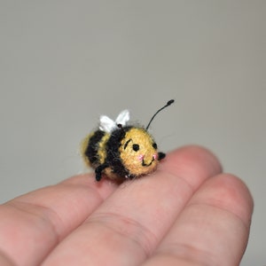 May include: A small crocheted bee with black and yellow stripes, white wings, and a cute embroidered face. The bee is sitting on a hand.