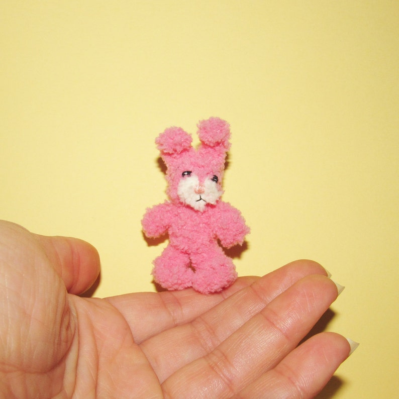 Puede incluir: Un peque&ntilde;o juguete de peluche de conejo rosa con rasgos faciales blancos. El conejo est&aacute; sentado en la palma de una mano.