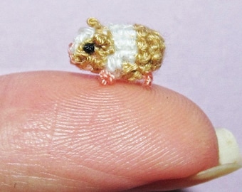 May include: A miniature crocheted guinea pig in shades of brown, white, and tan. The tiny animal has black eyes and pink feet. The small craft is displayed on a fingertip, showcasing its diminutive size. The background is a soft pink.