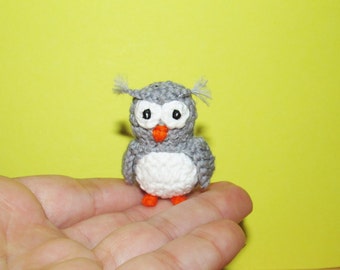 May include: A small gray crocheted owl with white eyes and an orange beak. The owl is sitting on a hand.