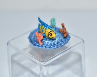 Puede incluir: Un pez de ganchillo amarillo y azul con una escena de arrecife de coral en un cubo de plástico transparente.