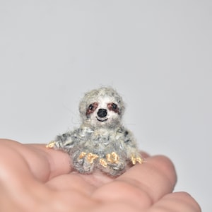 May include: A small, gray and white crocheted sloth with brown eyes and a black nose. The sloth is sitting on a hand.
