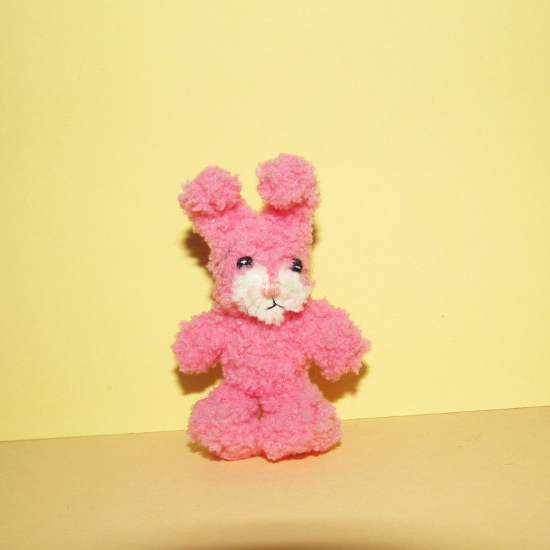 Puede incluir: Un juguete de conejo de peluche rosa con pelaje blanco alrededor de la nariz y ojos negros. El juguete est&aacute; de pie sobre una superficie amarilla.
