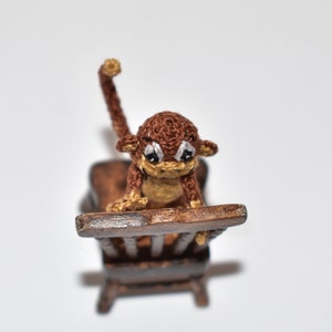 May include: A brown crocheted monkey doll sitting on a wooden chair. The monkey has a brown body, a tan face, and black eyes.