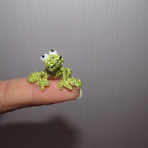 Puede incluir: Una pequeña rana de ganchillo hecha a mano, en tonos verdes, con ojos blancos y negros. La rana está posada en la punta de un dedo, mostrando sus delicados detalles y su diseño caprichoso. Un encantador ejemplo de arte de ganchillo.