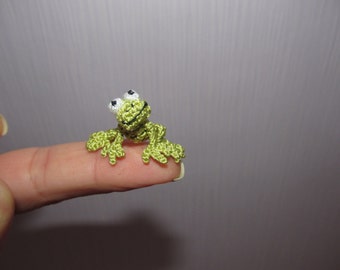 Op de afbeelding: Een kleine, handgemaakte gehaakte kikker in groentinten, met witte en zwarte ogen. De kikker zit op een vingertop, wat de delicate details en het grillige ontwerp laat zien. Dit miniatuuramfibie is een charmant voorbeeld van haakkunst.