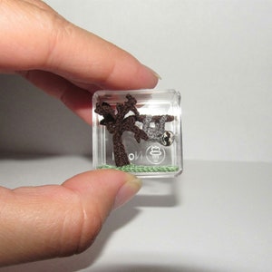 May include: A miniature crocheted sloth hanging from a brown tree branch in a clear plastic cube. The sloth is grey and white with black eyes. The tree is brown and has green leaves. The cube has a green base and a clear top.