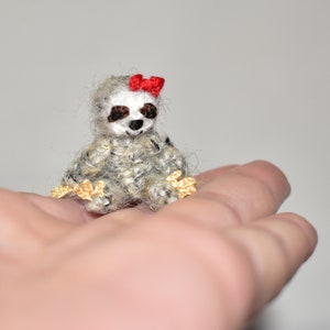 May include: A small crocheted sloth wearing a red bow, sitting on a hand. The sloth is gray and white with gold accents on its paws.