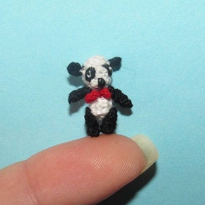 Puede incluir: Un pequeño oso panda de crochet con una pajarita roja. El panda es blanco con orejas, ojos y patas negras. Está de pie sobre un dedo.