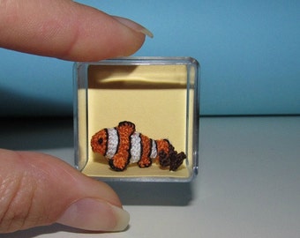 Puede incluir: Un pequeño pez payaso de ganchillo naranja y blanco en una vitrina de plástico transparente con fondo amarillo. La vitrina está sobre una superficie blanca.