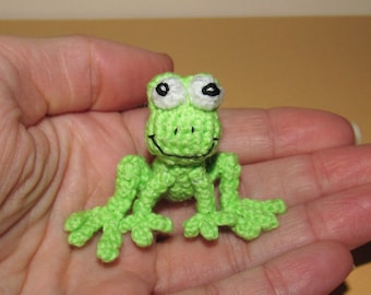 Op de afbeelding: Een kleine groene gehaakte kikker met witte ogen en zwarte pupillen. De kikker zit op een hand.