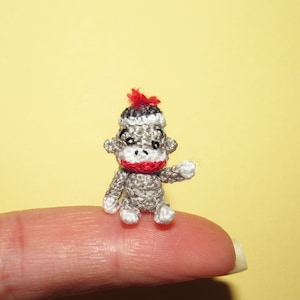 May include: A small crocheted monkey wearing a red and white hat. The monkey is sitting on a finger.