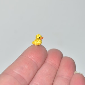 May include: A miniature, handmade yellow crochet duckling with an orange beak. The tiny duckling is displayed on a fingertip, emphasizing its small size and detailed craftsmanship. The background is a soft, neutral tone, contrasting with the bright yellow of the duck.