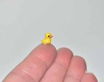 May include: A tiny, handmade yellow crochet duckling with an orange beak. The miniature duckling is perched on a fingertip, showcasing its small size and intricate details. The background is a soft, neutral color, highlighting the bright yellow of the duck.