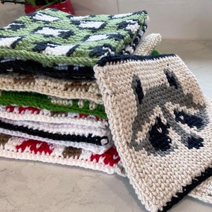 Könnte beinhalten: Ein Stapel handgefertigter Topflappen aus Häkelarbeit in verschiedenen Farben und Mustern. Ein Topflappen zeigt ein Waschbären-Design. Weitere Muster sind Grün und Weiß, Schwarz und Weiß sowie Rot und Braun. Die Topflappen sind auf einer hellen Oberfläche gestapelt.