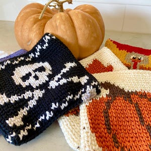 Puede incluir: Una colección de paños de cocina cuadrados de ganchillo con varios diseños, incluyendo una calavera y tibias cruzadas en blanco y negro, una calabaza naranja y un pavo. Los artículos están apilados con una calabaza naranja detrás.