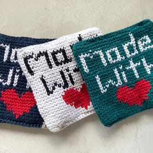 May include: Three crocheted squares in navy blue, white, and teal. Each square features the phrase "made with" in white, with a red heart below. The squares are handmade and likely used as kitchen accessories.