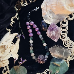May include: A multi-coloured crystal beaded necklace with amethyst, green, and clear beads. The necklace is displayed on a black velvet surface with other crystals and a skull.