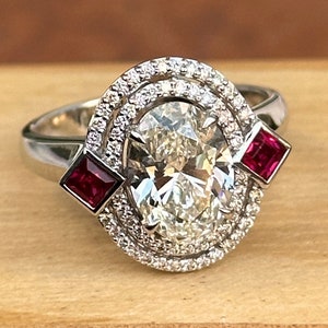 May include: A white gold engagement ring featuring a large oval-shaped diamond with a double halo of smaller diamonds. Two square-shaped ruby accent stones are set on either side of the diamond.