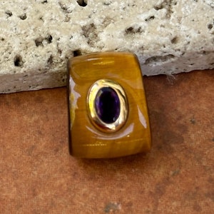 May include: A rectangular pendant with a gold setting and a dark purple oval gemstone. The pendant has a warm, amber-colored base with a polished finish. The gold setting surrounds the gemstone, creating a striking contrast.