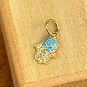 Könnte beinhalten: Goldener Anhänger in Form einer Hamsa-Hand mit einem blauen bösen Auge in der Mitte. Die Hamsa ist mit klaren, funkelnden Steinen verziert.