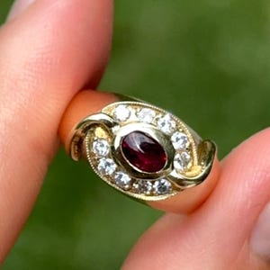 May include: A gold ring featuring a central oval-cut, deep red gemstone, surrounded by a halo of smaller, clear gemstones. The ring's band has an ornate, swirling design. The ring is held up against a blurred green background.
