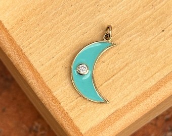Könnte beinhalten: Ein goldener Halbmond-Anhänger mit einem kleinen, klaren Edelstein in der Mitte. Der Mond ist in einem hellblauen Türkis lackiert.