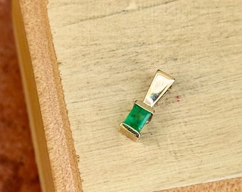 Könnte beinhalten: Ein kleiner, quadratischer Anhänger mit einem smaragdgrünen Edelstein und einer goldenen Fassung. Der Anhänger liegt auf einer Holzoberfläche.