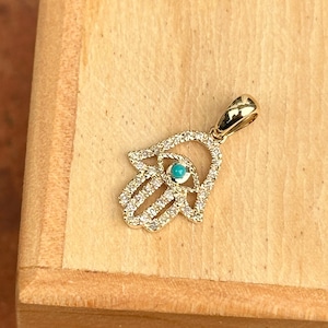 Könnte beinhalten: Goldfarbener Hamsa-Hand-Anhänger mit einem türkisfarbenen Mittelstein und klaren Kristallakzenten. Der Anhänger liegt auf einer Holzoberfläche. Die Hamsa-Hand ist ein Symbol für Schutz und Glück.