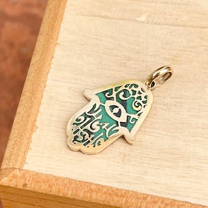 Könnte beinhalten: Goldener Hamsa-Hand-Anhänger mit grünem bösen Auge. Der Anhänger ist aufwendig verziert und hat ein filigranes Muster.